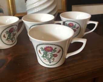 May include: Four white ceramic teacups with floral designs and green trim. The cups have a flared shape and a handle on the right side. The floral designs feature pink flowers, green leaves, and butterflies.