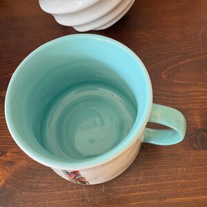 May include: A ceramic mug with a light blue interior and handle. The exterior is cream-colored with horizontal lines and floral accents. The mug sits on a dark wooden surface.