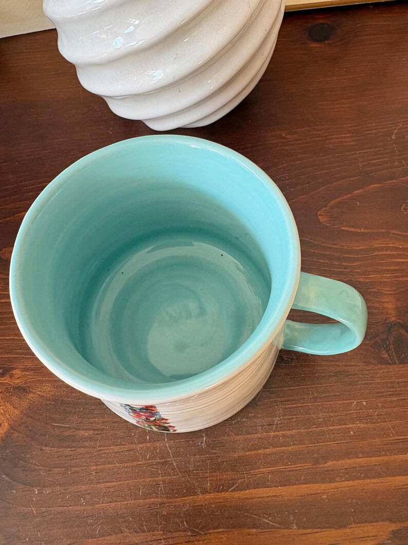 May include: A ceramic mug with a light blue interior and handle. The exterior is cream-colored with horizontal lines and floral accents. The mug sits on a dark wooden surface.