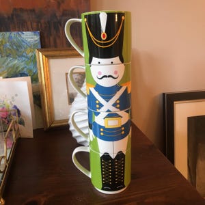 May include: A stack of four green and white ceramic mugs featuring a whimsical illustration of a toy soldier. The soldier is wearing a blue uniform with white stripes and a red and black hat.