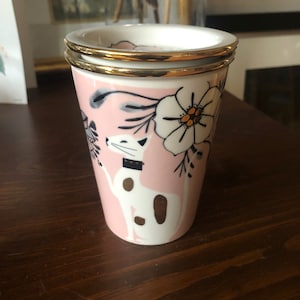 Op de afbeelding: Een roze keramische mok met een gouden rand en een witte kat met zwarte ogen en een gouden halsband. De kat wordt omringd door roze en witte bloemen met gouden accenten.