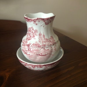 May include: A white ceramic jug with a red and white floral design. The jug has a scalloped rim and a wide base. It is sitting on a matching saucer.