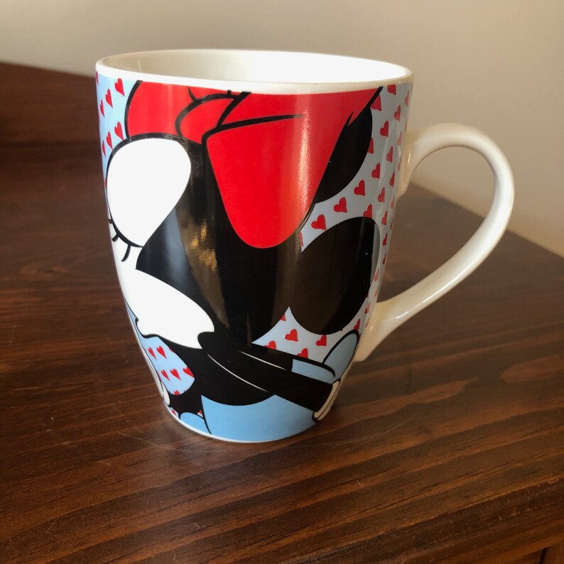 Pu&ograve; includere: Una tazza in ceramica bianca con un'immagine colorata di Minnie Mouse. La tazza ha un fiocco rosso, occhi neri e uno sfondo blu e bianco con cuori rossi.
