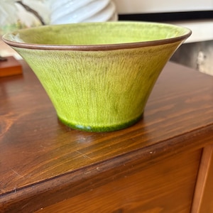 May include: A green ceramic bowl with a flared shape and a brown rim. The bowl has a textured surface and sits on a wooden table. The bowl is a decorative item.