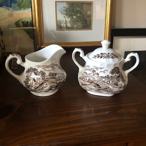 May include: A brown and white creamer and sugar set with a country scene design. The creamer and sugar are both made of ceramic and have a handle.