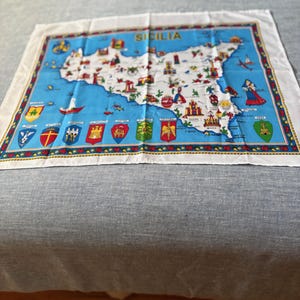 May include: A white cotton fabric square featuring a colourful map of Sicily, Italy. The map is bordered by a yellow trim with a gold chain design. The word "SICILIA" is printed in red at the top of the map. The map includes various landmarks and symbols, such as a church, a castle, a lighthouse, and a boat. The fabric also displays a row of colourful coats of arms at the bottom.