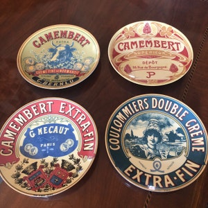 May include: Four vintage-style ceramic plates with different designs featuring the words "Camembert" and "Coulommiers Double Creme" in red, blue, and gold lettering. Each plate has a unique design with a cow, a woman, and a building.