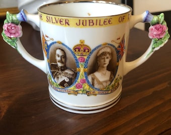 Puede incluir: Taza de cerámica blanca con dos asas decoradas con diseños florales. La taza presenta un retrato del rey Jorge V y la reina María con el texto "The Silver Jubilee of" alrededor de la parte superior.