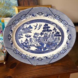 May include: An oval ceramic platter featuring a blue and white Willow pattern. The central scene depicts a pagoda, trees, a bridge, and two birds in flight. The wide border is decorated with a geometric lattice design and floral motifs.