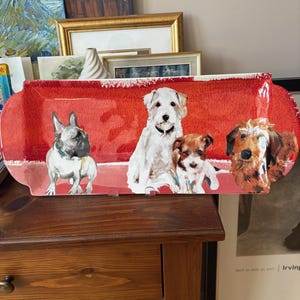 May include: A rectangular red ceramic serving platter featuring illustrations of four different dog breeds. The dogs are depicted in a cartoon style, with a French Bulldog, a white terrier, a small brown dog, and a terrier mix. The platter has a white border.