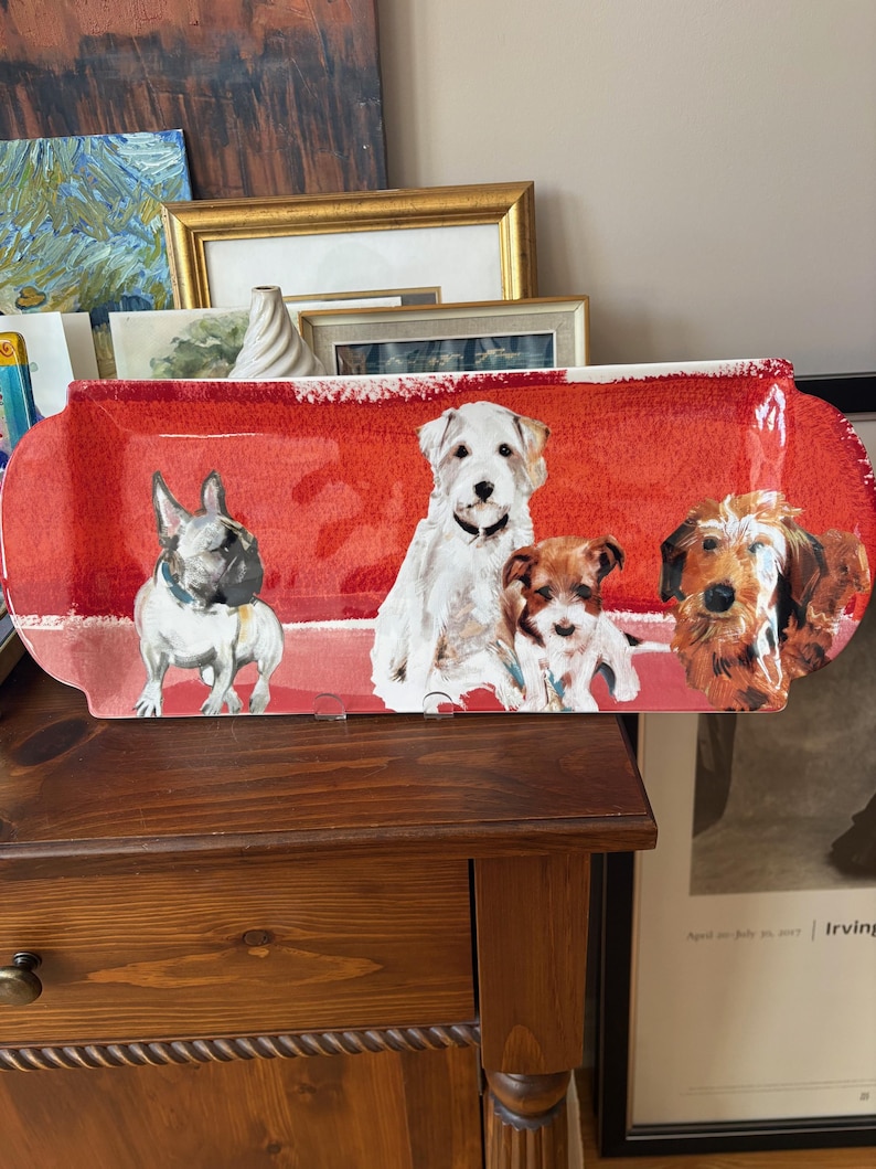 May include: A rectangular red ceramic serving platter featuring illustrations of four different dog breeds. The dogs are depicted in a cartoon style, with a French Bulldog, a white terrier, a small brown dog, and a terrier mix. The platter has a white border.