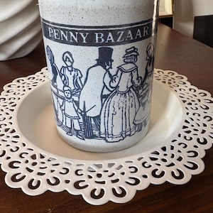 May include: A ceramic tumbler with a speckled white and brown finish. The tumbler features a blue illustration of people and the text "PENNY BAZAAR". It sits on a white, lacy, circular plate. A decorative item for the home.