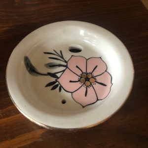 Op de afbeelding: Een witte keramische schotel met een gouden rand en een roze bloem met zwarte en gouden details. De schotel heeft twee kleine gaatjes in het midden.