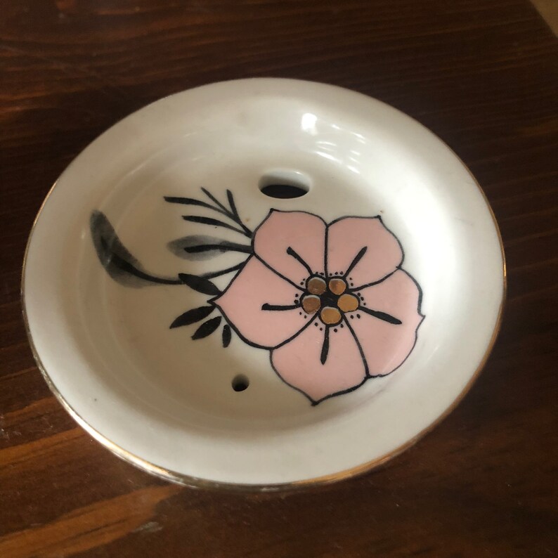 Op de afbeelding: Een witte keramische schotel met een gouden rand en een roze bloem met zwarte en gouden details. De schotel heeft twee kleine gaatjes in het midden.