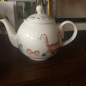 May include: White ceramic teapot with a cat design. The teapot has a lid with the words "Puss in Boots" written on it. The cats are wearing blue boots.