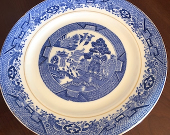 May include: A blue and white porcelain plate with a gold rim. The plate features a traditional willow pattern with a pagoda, a bridge, and a willow tree.