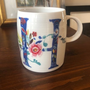 K&ouml;nnte beinhalten: Wei&szlig;e Keramiktasse mit blauem und floralem Design mit dem Buchstaben "H".