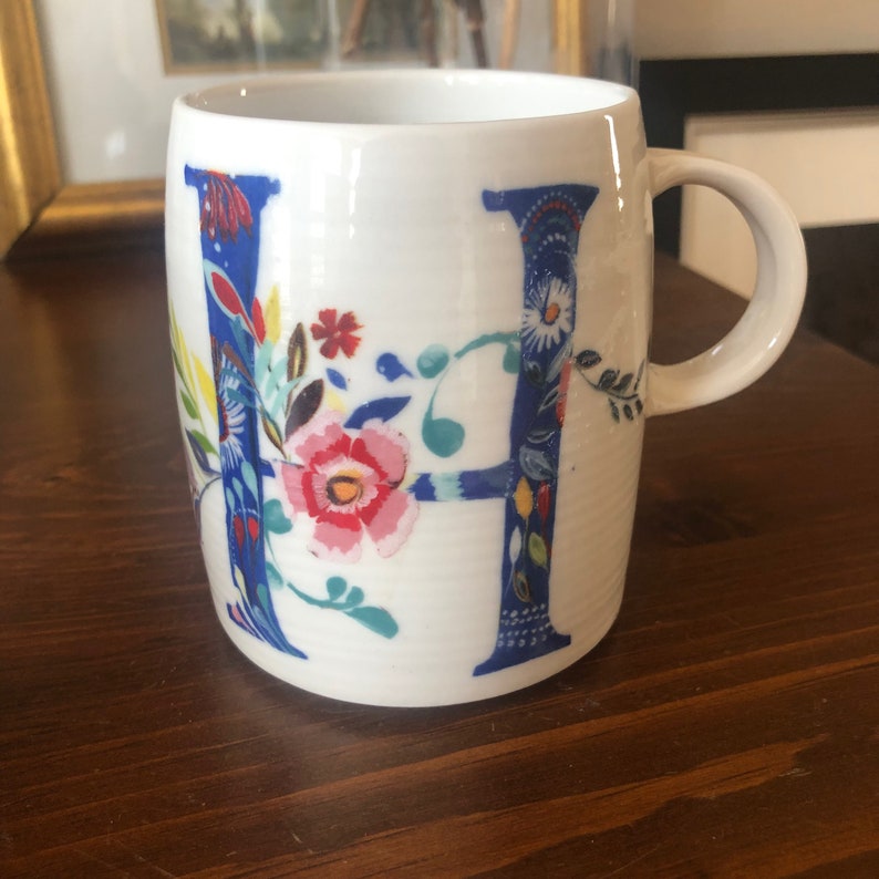 K&ouml;nnte beinhalten: Wei&szlig;e Keramiktasse mit blauem und floralem Design mit dem Buchstaben "H".