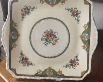 May include: A white square plate with a scalloped edge and a floral design. The plate has a green and gold border with a blue and gold inner border. The center of the plate has a floral design with a green and gold border.