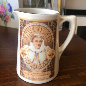 May include: Cream-colored ceramic pitcher with a handle, featuring a vintage "White Queen Whisky" label. The label includes an illustration of a woman, a plaid background, and the text "William Dyson & Co. Manchester."