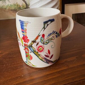 K&ouml;nnte beinhalten: Wei&szlig;e Keramiktasse mit einem bunten Blumenmuster und dem Buchstaben "K" in der Mitte. Die Tasse hat einen Henkel und steht auf einer Holzoberfl&auml;che.