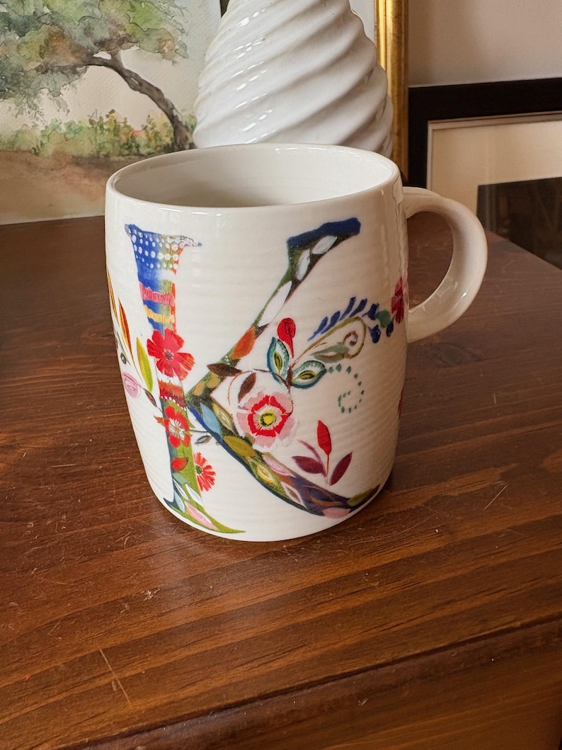 K&ouml;nnte beinhalten: Wei&szlig;e Keramiktasse mit einem bunten Blumenmuster und dem Buchstaben "K" in der Mitte. Die Tasse hat einen Henkel und steht auf einer Holzoberfl&auml;che.