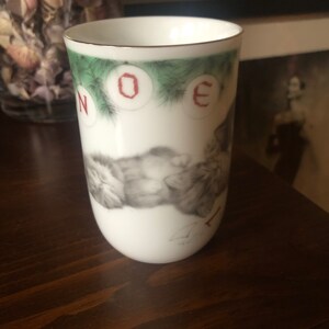 May include: White ceramic mug with a gold rim and a Christmas design. The mug features a picture of two cats sleeping on a bed of pine needles with red ornaments that spell out "NOEL" on the top.