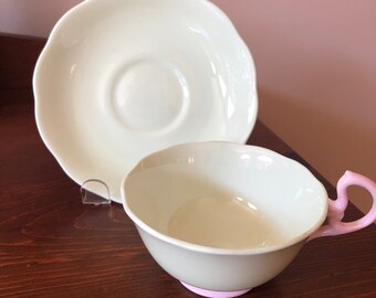May include: A white teacup and saucer set with a pink rim and handle. The saucer has a scalloped edge.