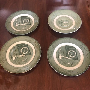 May include: Set of four green and white ceramic plates with a vintage design featuring a spinning wheel and other tools. The plates have a raised rim and a decorative border.