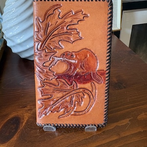 May include: A rectangular tan leather wallet featuring a hand-tooled design of a beaver holding a branch surrounded by large maple leaves. The edges of the wallet are finished with a dark brown braided leather lacing detail throughout the perimeter.