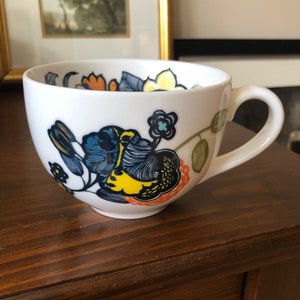 Pu&ograve; includere: Tazza in ceramica bianca con un design floreale in tonalit&agrave; di blu, giallo, arancione e verde. I fiori sono stilizzati e hanno un aspetto fantasioso.