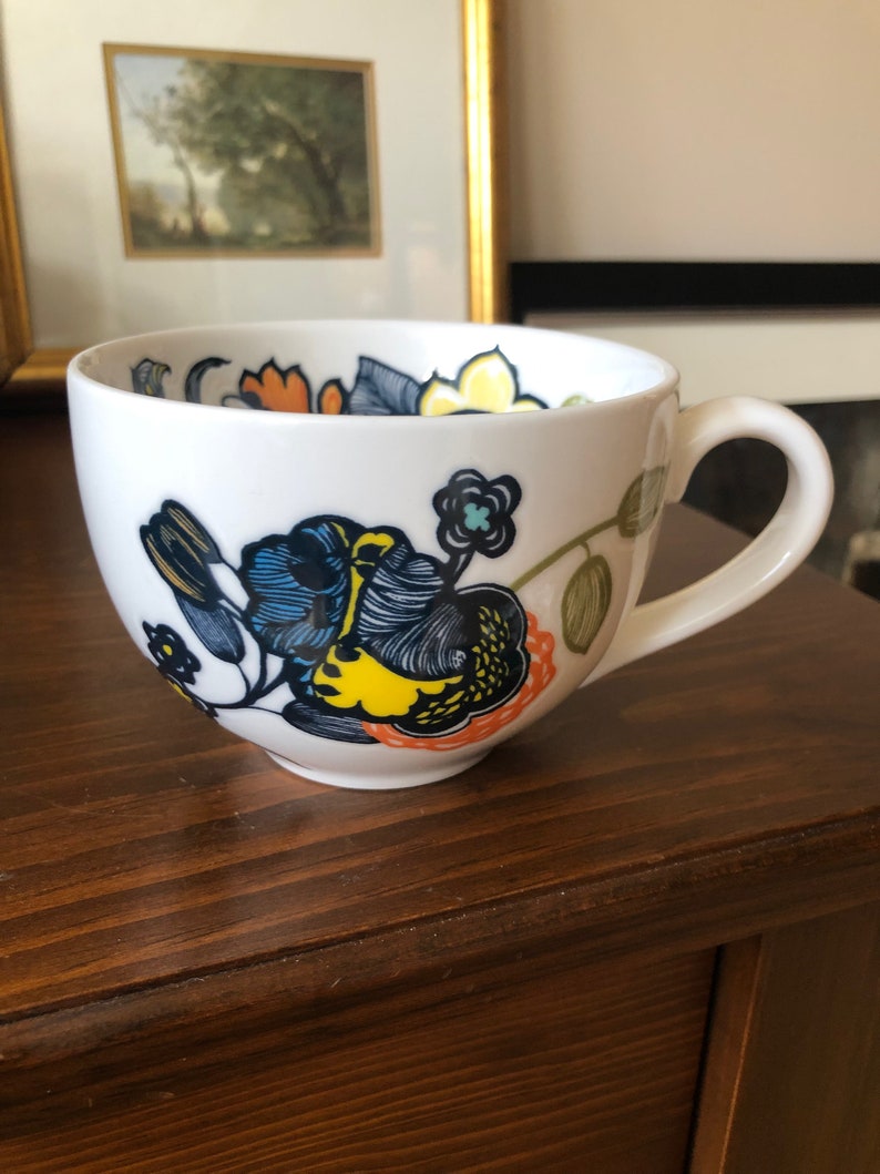 Pu&ograve; includere: Tazza in ceramica bianca con un design floreale in tonalit&agrave; di blu, giallo, arancione e verde. I fiori sono stilizzati e hanno un aspetto fantasioso.