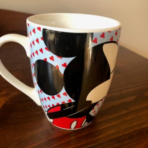 Pu&ograve; includere: Tazza in ceramica bianca con sfondo blu e bianco con un'immagine in bianco e nero di Topolino con cuori rossi. La tazza ha un manico bianco.