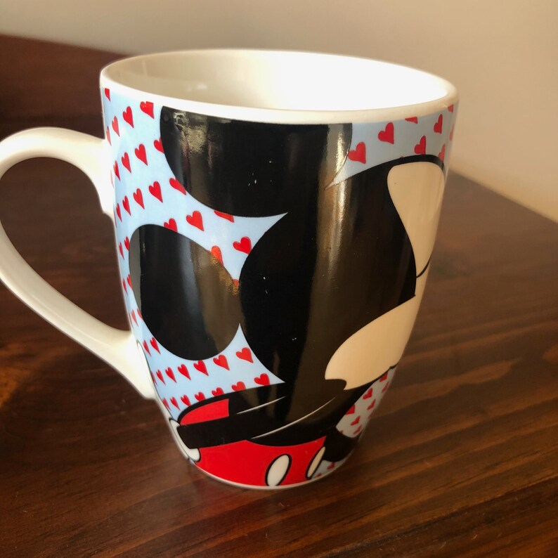 Pu&ograve; includere: Tazza in ceramica bianca con sfondo blu e bianco con un'immagine in bianco e nero di Topolino con cuori rossi. La tazza ha un manico bianco.