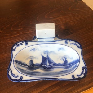 May include: A blue and white porcelain soap dish with a raised handle. The dish features a scene of sailing ships and a windmill in a seascape.