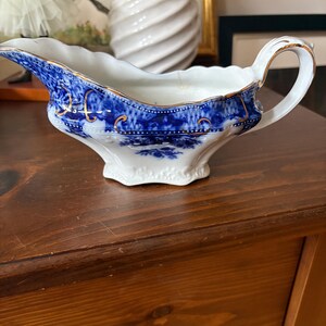 May include: A white and blue porcelain gravy boat with a handle and spout. The boat features a blue floral pattern with gold trim along the edges. The gravy boat is sitting on a wooden surface.
