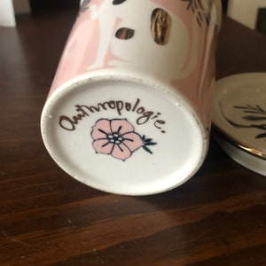 Op de afbeelding: De onderkant van een roze en witte keramische mok met gouden accenten. De mok heeft een roze bloemenontwerp en de tekst "Anthropologie."