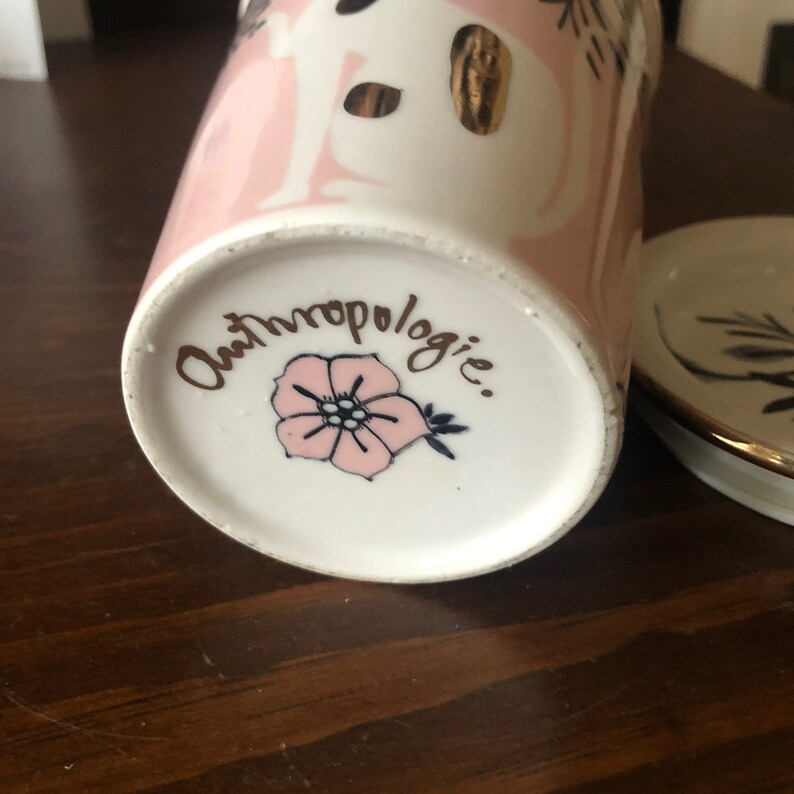 Op de afbeelding: De onderkant van een roze en witte keramische mok met gouden accenten. De mok heeft een roze bloemenontwerp en de tekst "Anthropologie."
