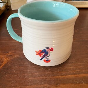 May include: White ceramic mug with a light blue interior and handle. The mug features a bird and floral design in shades of blue, red, and orange. The mug has a ribbed texture and is suitable for hot or cold beverages.