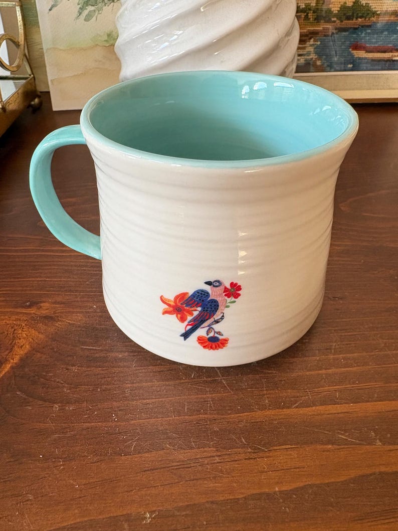 May include: White ceramic mug with a light blue interior and handle. The mug features a bird and floral design in shades of blue, red, and orange. The mug has a ribbed texture and is suitable for hot or cold beverages.