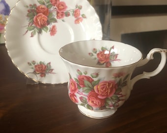 May include: A white teacup and saucer with a gold trim and a floral pattern of pink roses.