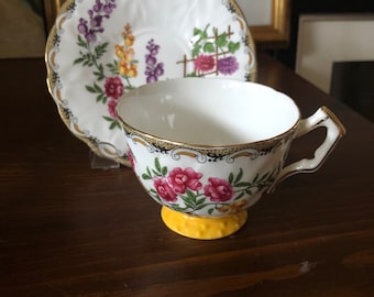 May include: A white teacup and saucer with a floral pattern. The teacup has a yellow base and the saucer has a gold rim.
