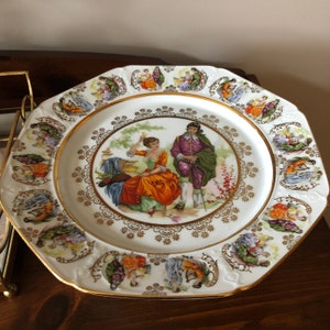 May include: A white porcelain plate with a gold rim and a central image of a couple in a garden setting. The plate is decorated with a border of smaller images of couples in various poses.
