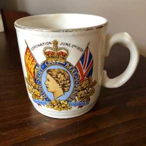 May include: White ceramic mug with a gold rim and a colourful image of Queen Elizabeth II's coronation. The image includes the royal crown, a banner with the words "Dieu et mon Droit", and the text "Coronation June 2nd 1953".