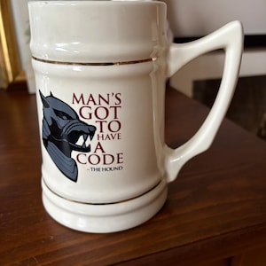 May include: White ceramic mug with a gold rim and a black and gray wolf graphic. The mug has the text "Man's Got To Have A Code - The Hound" printed on it.