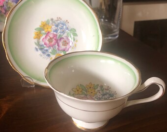 May include: A white teacup and saucer with a green rim and floral pattern. The floral pattern features pink, yellow, and blue flowers.