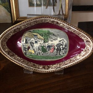 Peut inclure: Un plat ovale de couleur bordeaux avec un bord doré et une scène blanche de personnes dansant devant un chalet. Le plat a une bordure décorative avec des accents dorés.