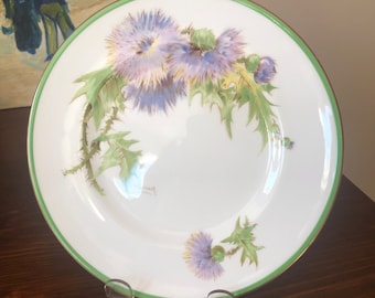 May include: A white porcelain plate with a green rim and a gold accent. The plate features a hand-painted design of purple and blue thistles with green leaves.