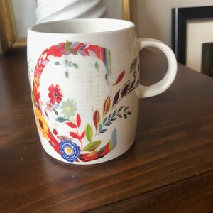 K&ouml;nnte beinhalten: Wei&szlig;e Keramiktasse mit einem Blumendesign, das einen gro&szlig;en Buchstaben C in Rot, Gelb, Blau, Gr&uuml;n und Orange zeigt. Die Tasse hat einen Henkel.