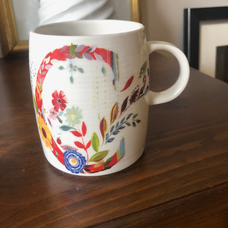 K&ouml;nnte beinhalten: Wei&szlig;e Keramiktasse mit einem Blumendesign, das einen gro&szlig;en Buchstaben C in Rot, Gelb, Blau, Gr&uuml;n und Orange zeigt. Die Tasse hat einen Henkel.